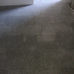 Ułożona podłoga z szarych, prostokątnych płytek w trakcie remontu, widoczne białe płytki na ścianie i fragment przejścia do innego pomieszczenia.