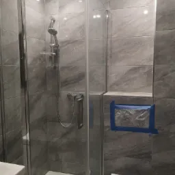 Szklana kabina prysznicowa z szarymi płytkami imitującymi marmur w łazience w Nowym Tomyślu, z widocznym zabezpieczeniem folią i taśmą malarską miejsca na półkę.