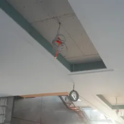 Niedokończony sufit z wnękami, widoczne przewody elektryczne, konstrukcja z płyt w otworach, częściowo pomalowany na biało, drabina w tle.