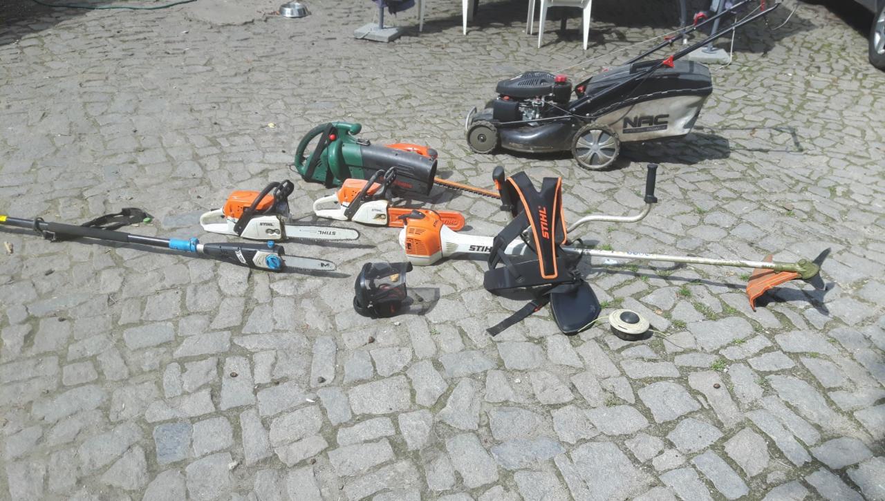 Zestaw narzędzi ogrodniczych, w tym kosiarka NAC, dmuchawa, kilka pilarek łańcuchowych Stihl, podkaszarka z osprzętem i maska ochronna, ułożone na brukowanej powierzchni.