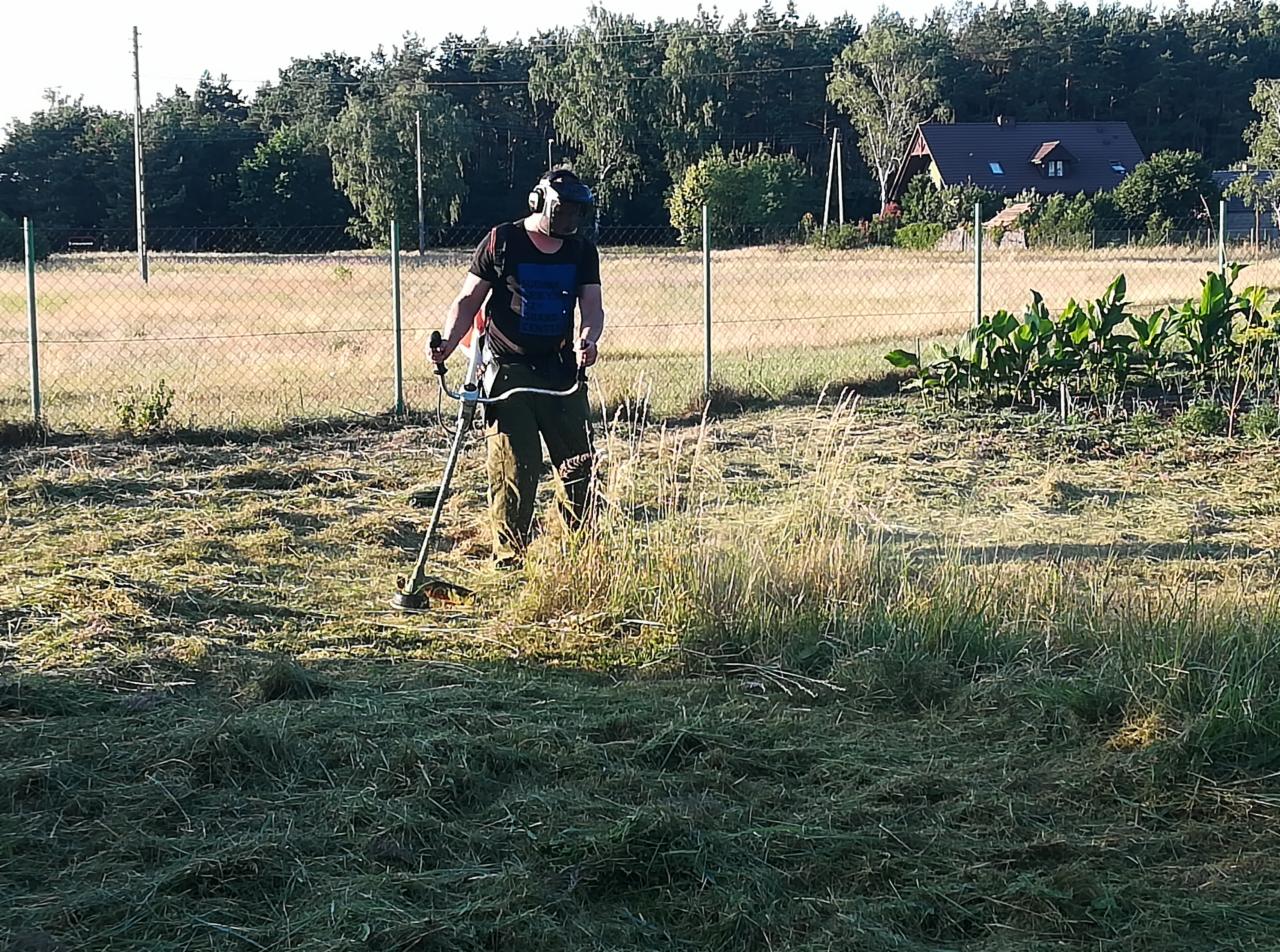 Pracownik w Gorzowie Wielkopolskim kosi trawę kosiarką spalinową na nierównym terenie, w tle widoczny płot z siatki i zabudowania.