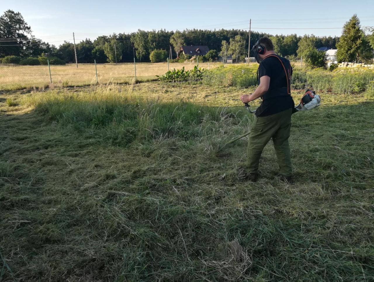 Mężczyzna w ochronnych słuchawkach kosi trawę na dużej, zarośniętej łące przy użyciu podkaszarki spalinowej Stihl, widoczne częściowo skoszone źdźbła trawy.