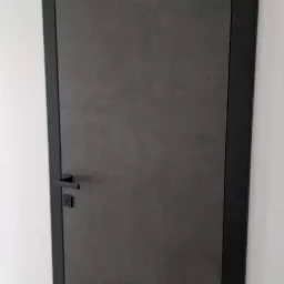 Matowe, grafitowe skrzydło drzwiowe z czarną ościeżnicą i minimalistyczną klamką na tle białej ściany.