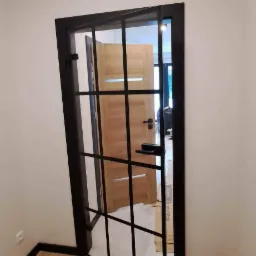 Szklane drzwi wewnętrzne z czarną ramą i podziałami na kwadraty, widok na drewniane drzwi w głębi pomieszczenia i drewnianą podłogę.