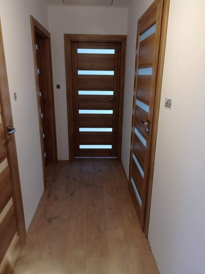 Korytarz z trzema drewnianymi drzwiami z poziomymi przeszkleniami, widoczna podłoga z paneli w odcieniach brązu, ściany w kolorze złamanej bieli, oświetlenie górne.