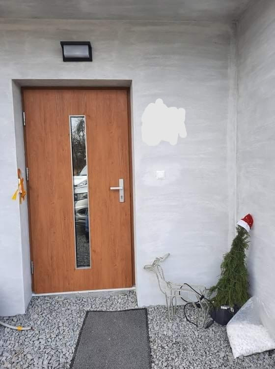 Zewnętrzne drzwi wejściowe z szybą, ozdobione świątecznym drzewkiem w czerwonej czapce i metalowym reniferem na tle szarego, kamienistego podłoża.