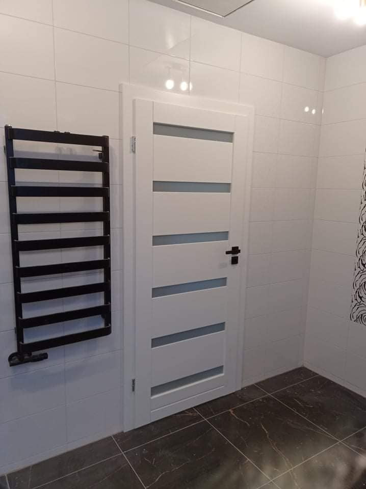 Białe drzwi wewnętrzne z poziomymi przeszkleniami w łazience z czarnym grzejnikiem drabinkowym i ciemną, marmurkową podłogą.