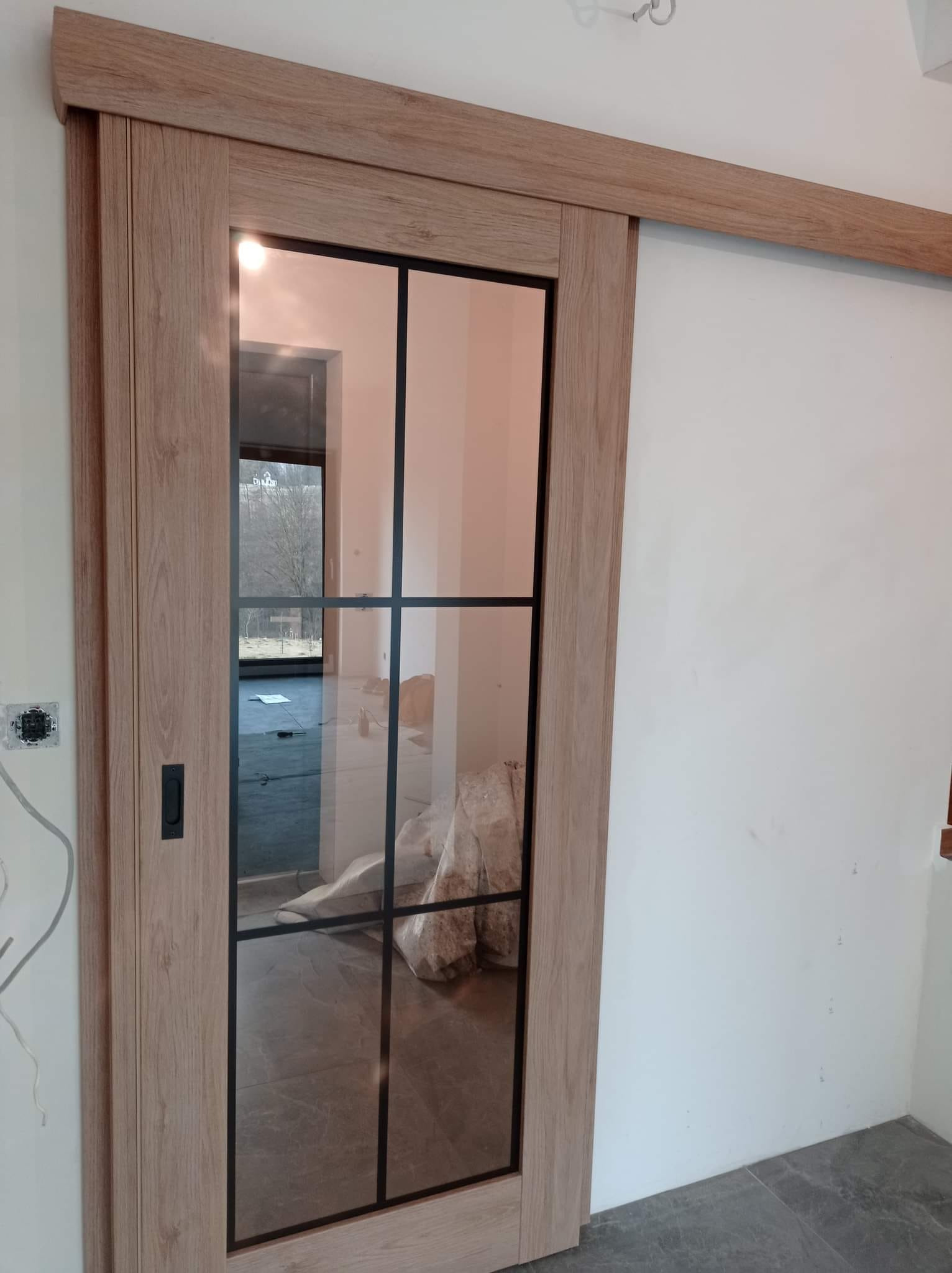 Szklane drzwi przesuwne w drewnianej ramie z czarnymi szprosami, widok z boku, widoczne niedokończone wykończenie wnętrza.