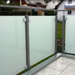 Nowoczesna zabudowa balkonu z matowego szkła i szarych metalowych słupków, widok z zewnątrz na tle domów jednorodzinnych.