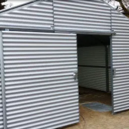 Metalowa konstrukcja z falistej blachy z częściowo otwartymi przesuwnymi drzwiami, widoczne mocowania szyn i zamek.