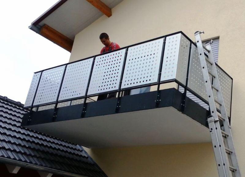 Montaż balustrady balkonowej z perforowanych paneli na nowo wybudowanym balkonie, widoczna drabina oparta o ścianę budynku.