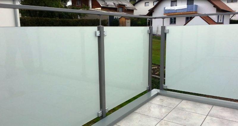 Nowoczesna zabudowa balkonu z matowego szkła i szarych metalowych słupków, widok z zewnątrz na tle domów jednorodzinnych.