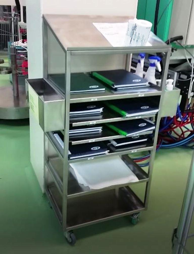 Wielopoziomowy wózek ze stali nierdzewnej na kółkach, z przegródkami wypełnionymi notatnikami, środkami czyszczącymi i papierem, stojący w pomieszczeniu o zielonej podłodze.