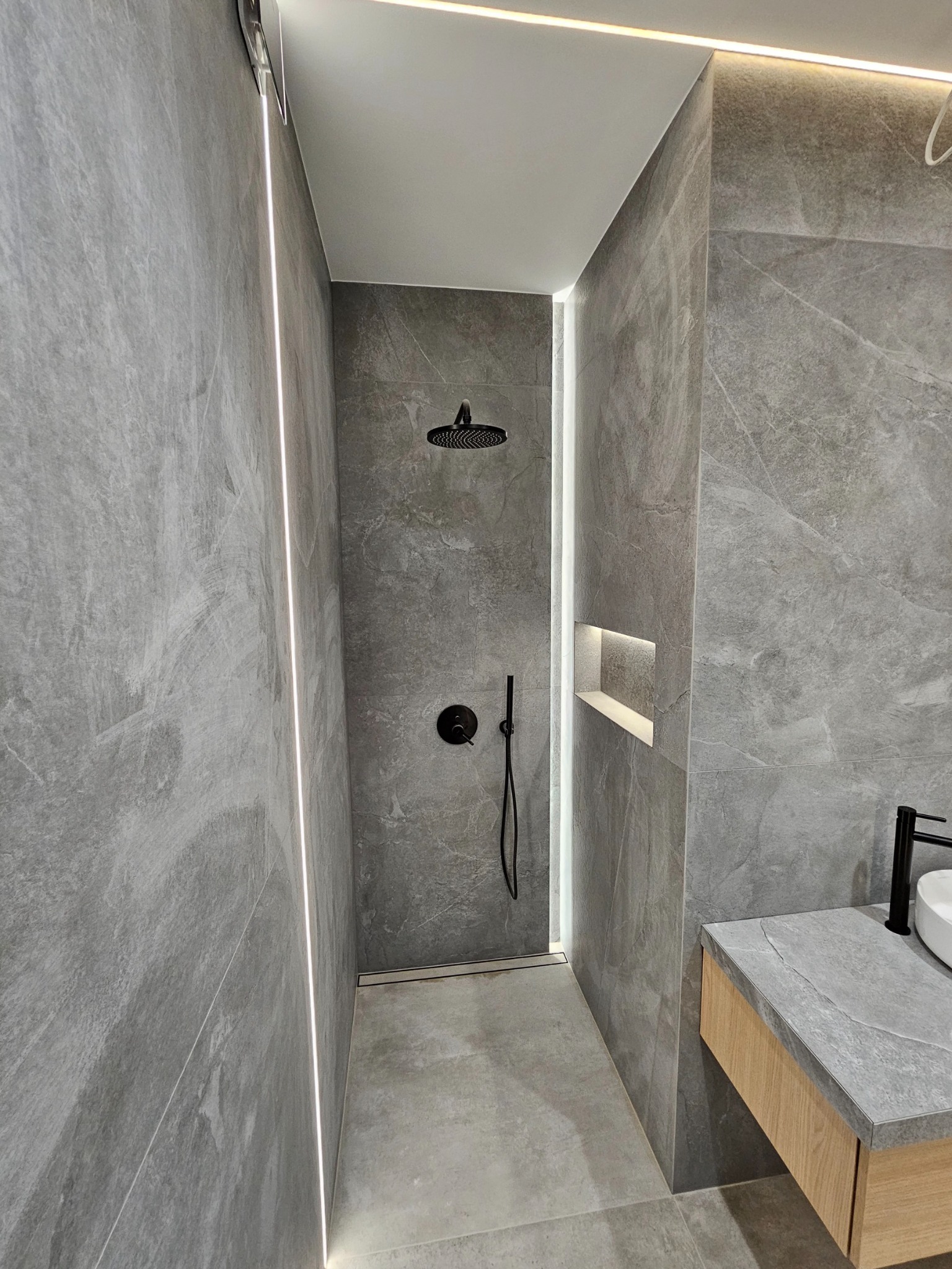 Nowoczesna kabina prysznicowa z szarymi płytkami imitującymi kamień, czarną armaturą i subtelnym oświetleniem LED wzdłuż krawędzi. Minimalistyczny design z wnęką na kosmetyki.