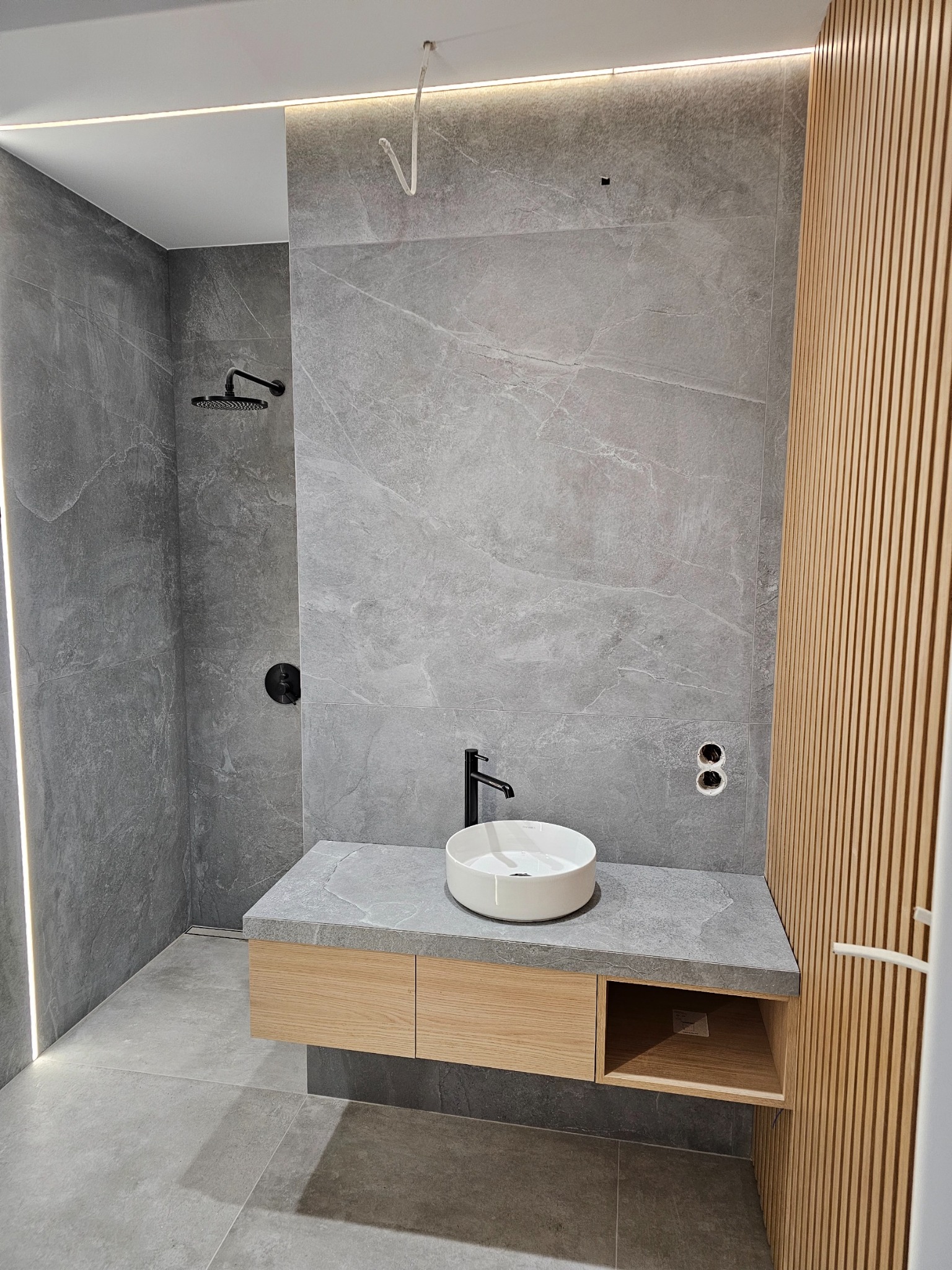 Nowoczesna łazienka z szarymi płytkami imitującymi marmur, drewnianą szafką pod umywalką i dekoracyjnymi lamelami. Minimalistyczny design z czarną armaturą.