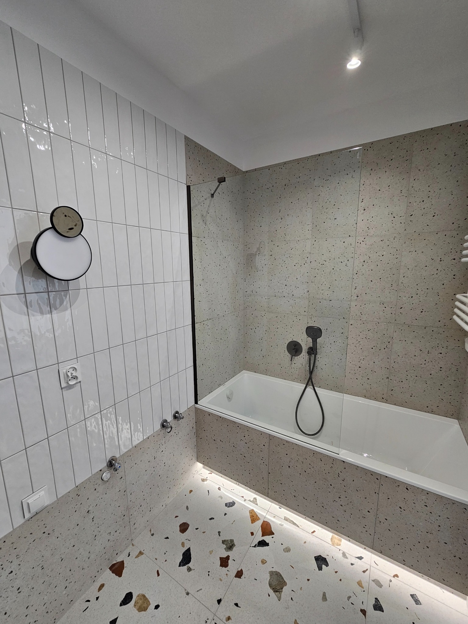 Nowoczesna łazienka z wanną, szklanym parawanem i oryginalnymi płytkami lastryko na podłodze i ścianach. Białe, pionowe płytki na ścianie z dekoracyjnym oświetleniem.