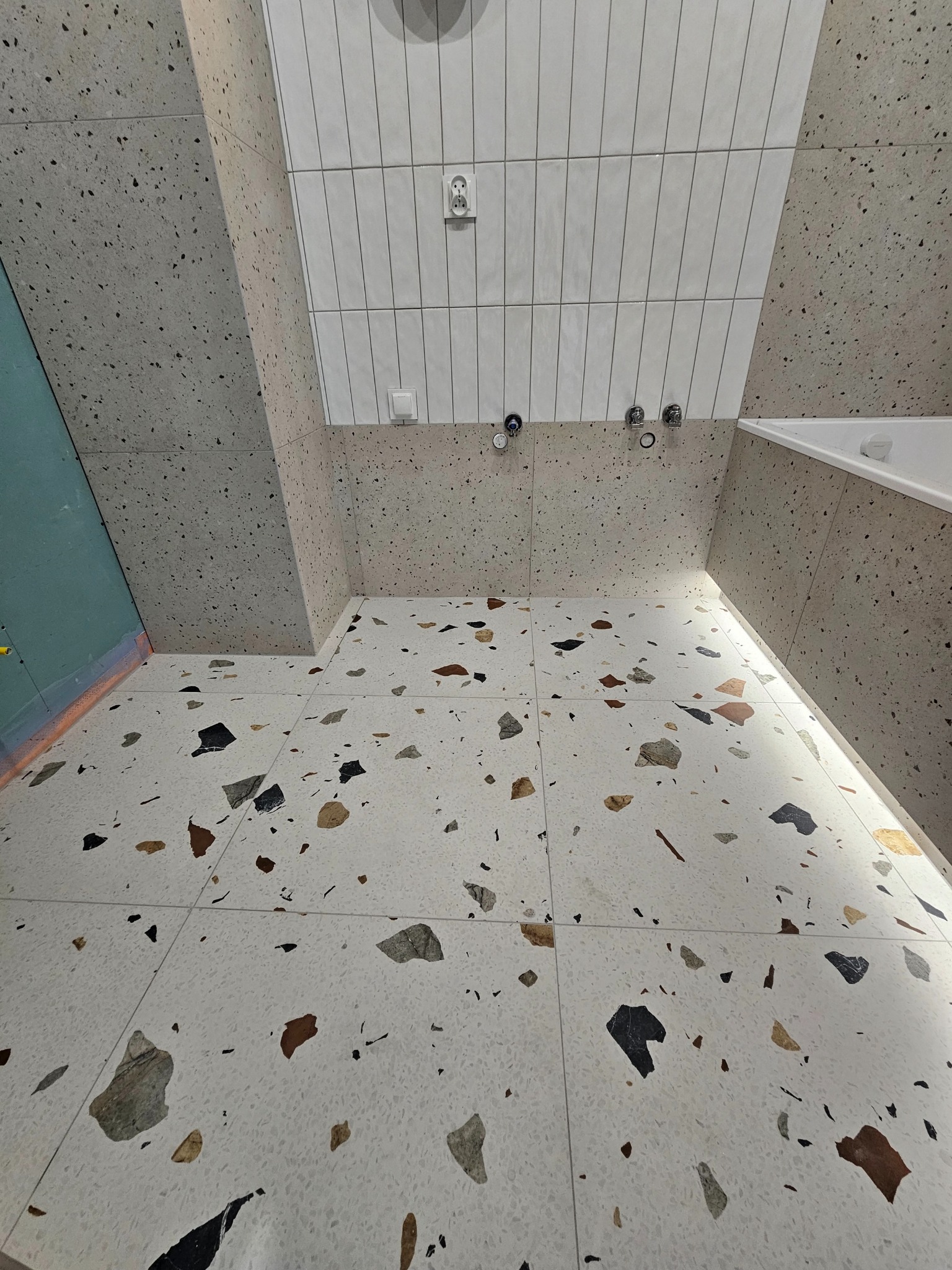 Wykończona łazienka z terazzo na podłodze i ścianach, białe płytki na górze. Widoczne baterie i fragment wanny. Ujęcie z góry podkreśla wzór.