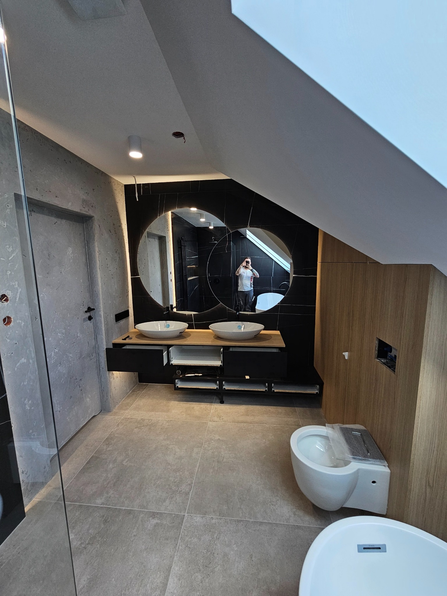 Nowoczesna łazienka na poddaszu z okrągłym lustrem, umywalkami nablatowymi i czarnymi płytkami. Widoczna toaleta i wanna. Ściany wykończone w szarości i drewnie.