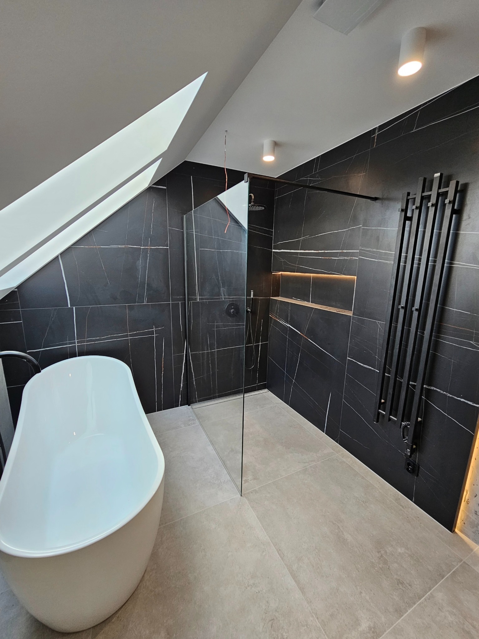 Nowoczesna łazienka na poddaszu z czarnymi płytkami imitującymi marmur, białą wanną wolnostojącą i grzejnikiem drabinkowym w kolorze czarnym.