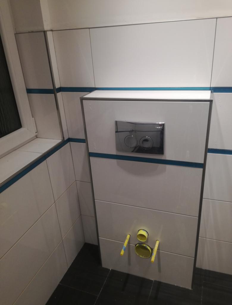 Biała zabudowa stelaża WC z chromowanym przyciskiem spłukującym i widocznymi rurami odpływowymi, wykończona białymi płytkami z niebieskim akcentem, na tle czarnej podłogi.