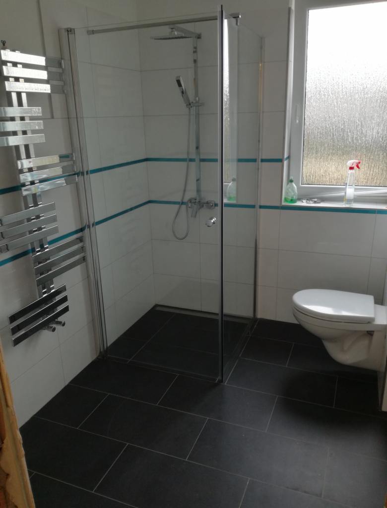 Wykończona łazienka z kabiną prysznicową typu walk-in, grzejnikiem drabinkowym, czarną podłogą z płytek i toaletą, wykończona białymi płytkami z turkusowym akcentem.