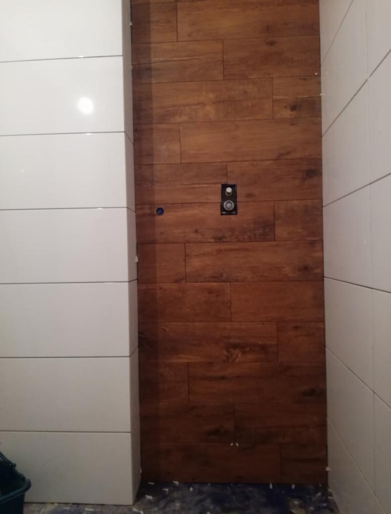 Ściana w łazience wyłożona białymi i drewnopodobnymi płytkami, widoczne gniazdko elektryczne i otwór na rurę, fuga jeszcze nieoczyszczona.