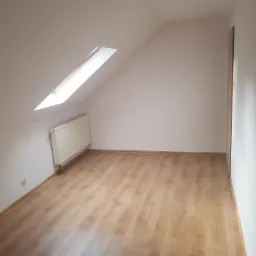 Puste, jasne poddasze z oknem dachowym, nowymi panelami podłogowymi w odcieniach drewna i białymi ścianami z grzejnikiem.