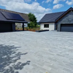 Szeroki podjazd z szarej kostki brukowej prowadzący do dwóch nowoczesnych domów z ciemnymi dachami i garażami, jeden z panelami słonecznymi, w tle zieleń i błękitne niebo z chmurami.