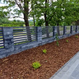 Nowoczesne ogrodzenie panelowe z elementami betonowymi, otoczone korą drzewną i młodymi roślinami, z fragmentem chodnika z kostki brukowej po prawej stronie, na tle zieleni drzew.