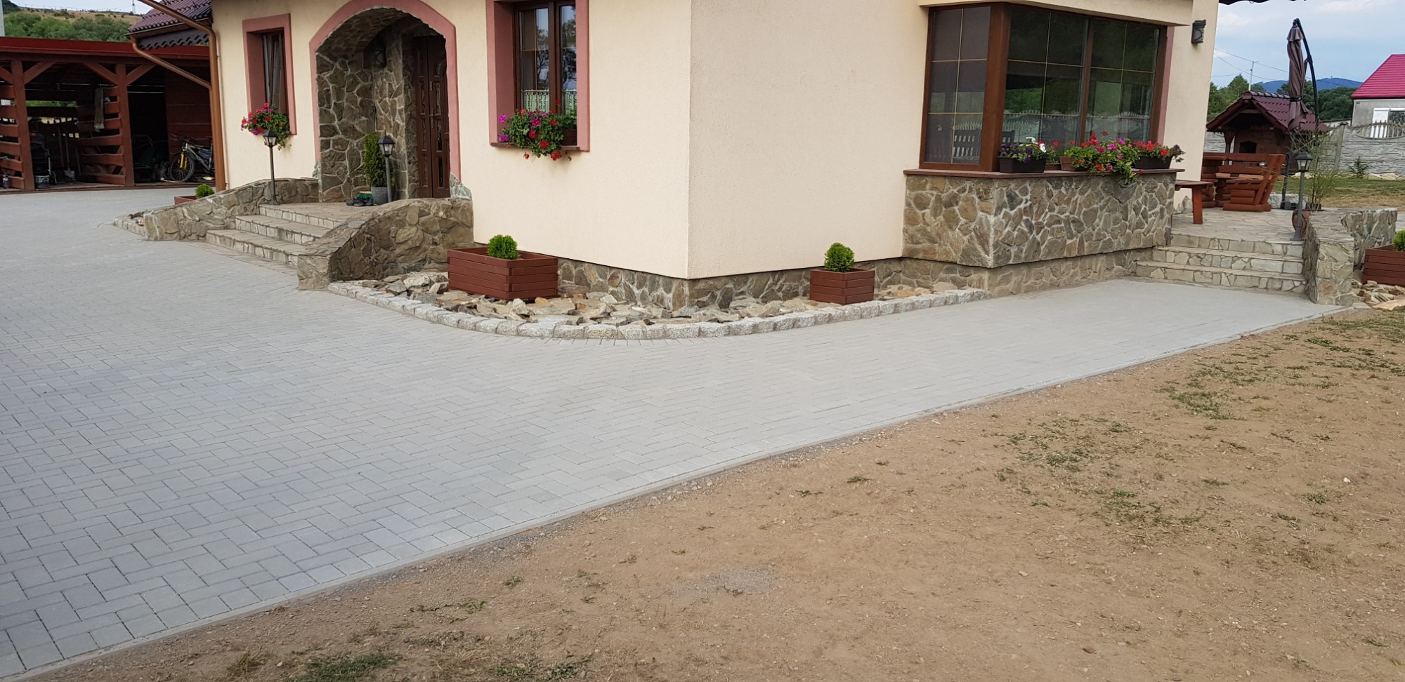 Ułożona szara kostka brukowa przed domem z kamienną elewacją, widoczne schody z kamienia i rośliny w donicach.