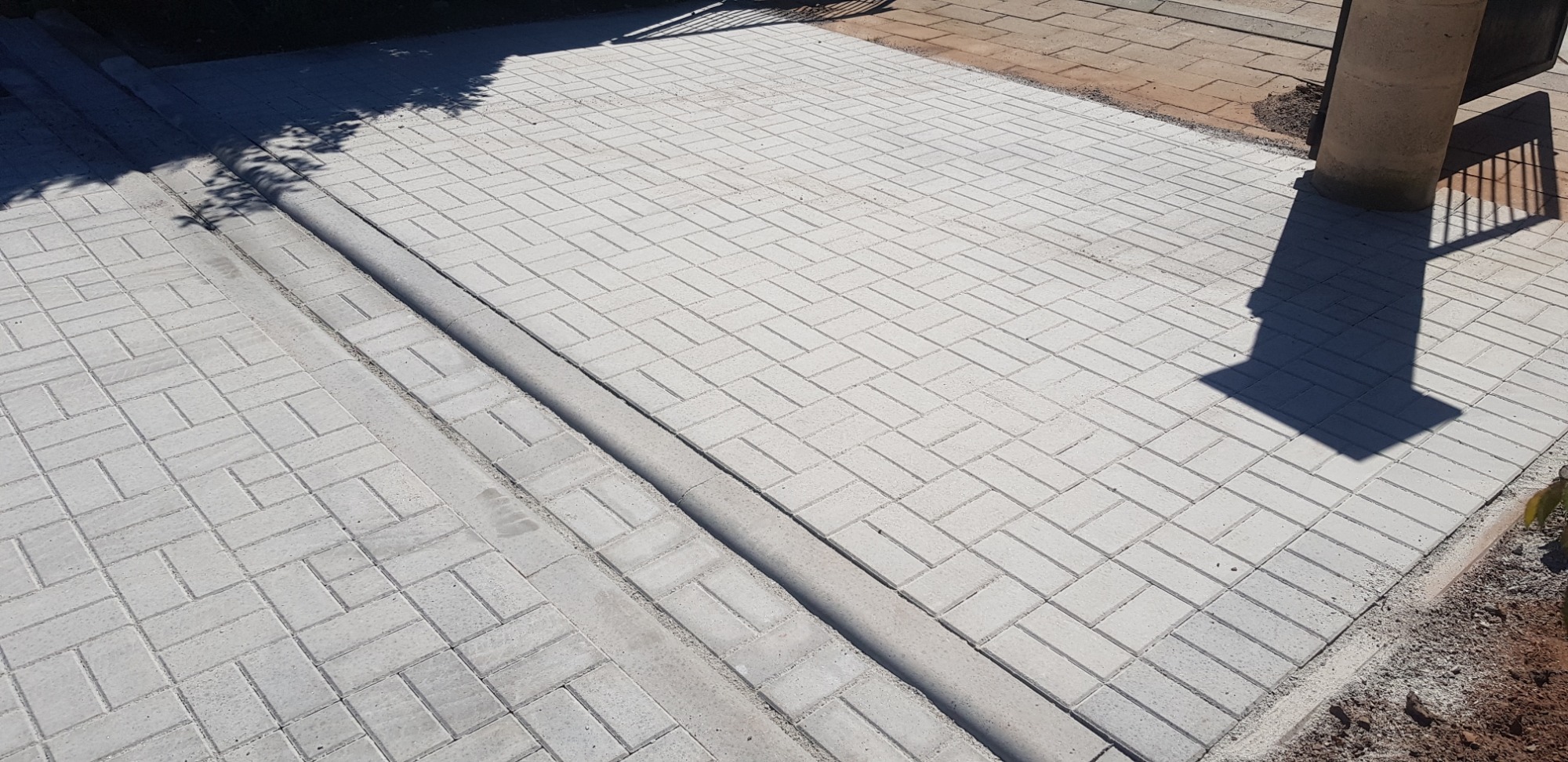 Nowo ułożona, jasnoszara kostka brukowa w wzór jodełki, z widocznym betonowym obrzeżem i cieniem drzewa, tuż po zakończeniu prac.