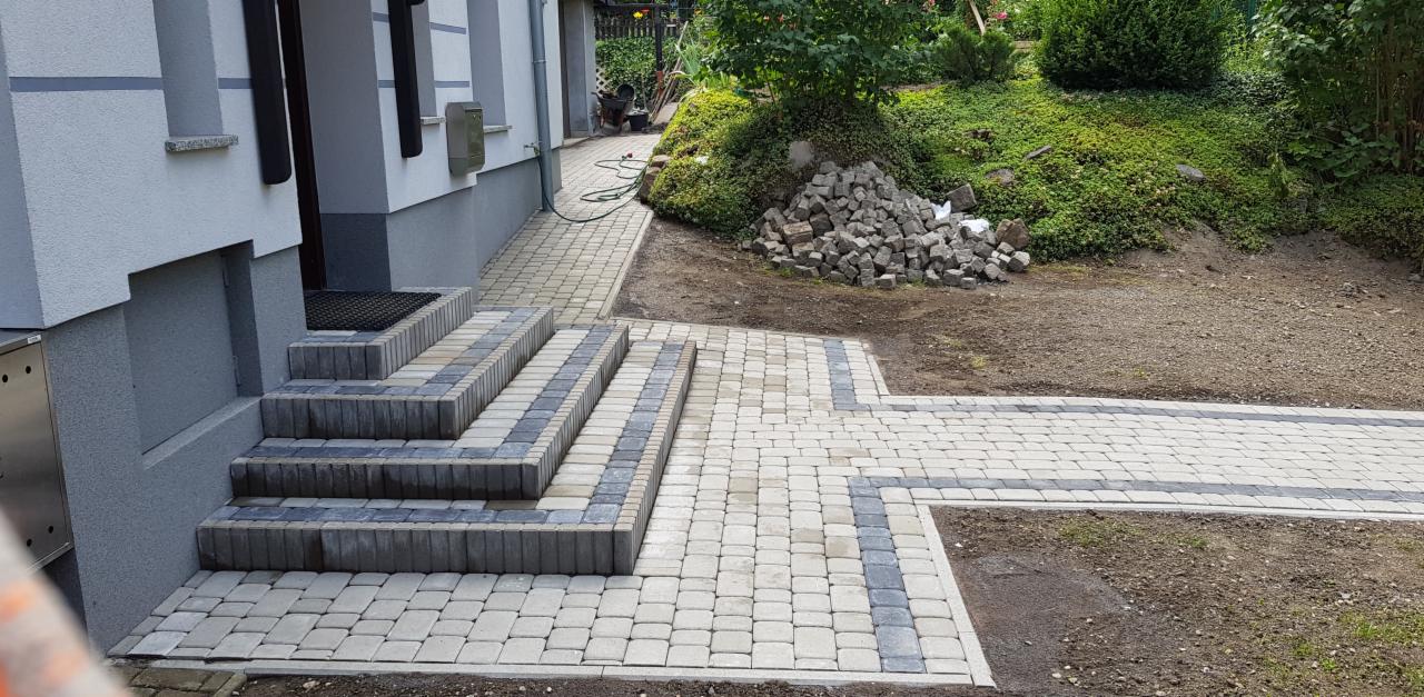 Ułożona kostka brukowa w Kamiennej Górze, tworząca schody wejściowe i ścieżkę, z użyciem dwóch odcieni szarości, prowadząca do domu z widocznym stosem kostki granitowej obok.