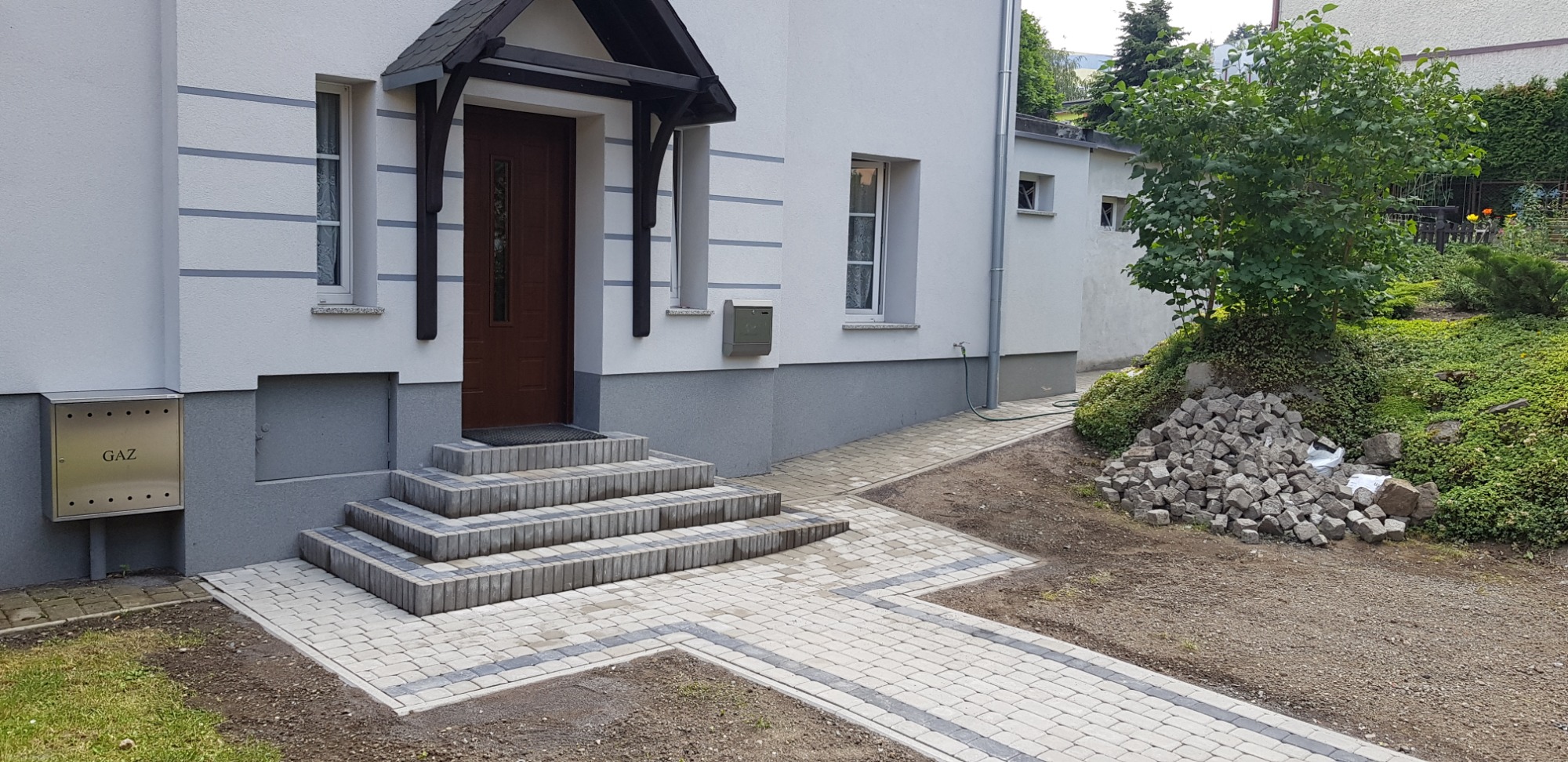 Nowo ułożona kostka brukowa przed domem z szarymi akcentami, schodami z kostki i stertą kostki granitowej obok rabaty z zielenią.