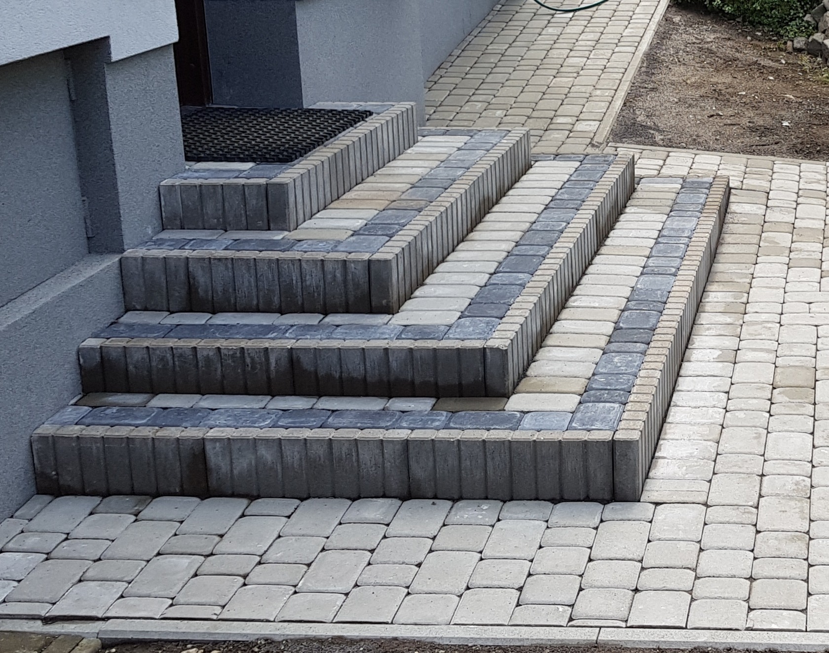 Schody wejściowe wyłożone kostką brukową w odcieniach szarości i beżu, dopasowane do chodnika z kostki, z widoczną wycieraczką przed drzwiami.