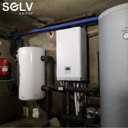 Zestaw urządzeń w kotłowni: wiszący biały piec gazowy Aquami Totenso z cyfrowym wyświetlaczem, obok biały zasobnik Galmet, rury w izolacji, na tle surowego betonu i logo firmy SELV Group w rogu.