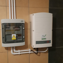 SELV GROUP - FOTOWOLTAIKA, POMPY CIEPŁA, KLIMATYZACJE - Skrzynka elektryczna z bezpiecznikami i falownik firmy SELV na kafelkowanej ścianie. Białe rury PCV prowadzą kable.