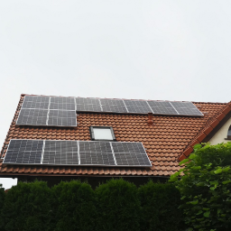 SELV GROUP - FOTOWOLTAIKA, POMPY CIEPŁA, KLIMATYZACJE - Panele słoneczne na dachu domu z czerwoną dachówką, widoczne krzewy i logo firmy na górze kadru.