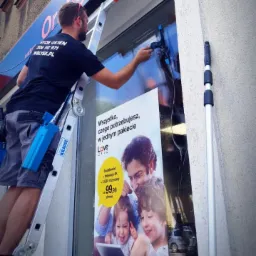 Pracownik myje okno na wysokości przy użyciu ściągaczki, stojąc na drabinie Krause. Na oknie naklejony plakat reklamowy sieci Orange.