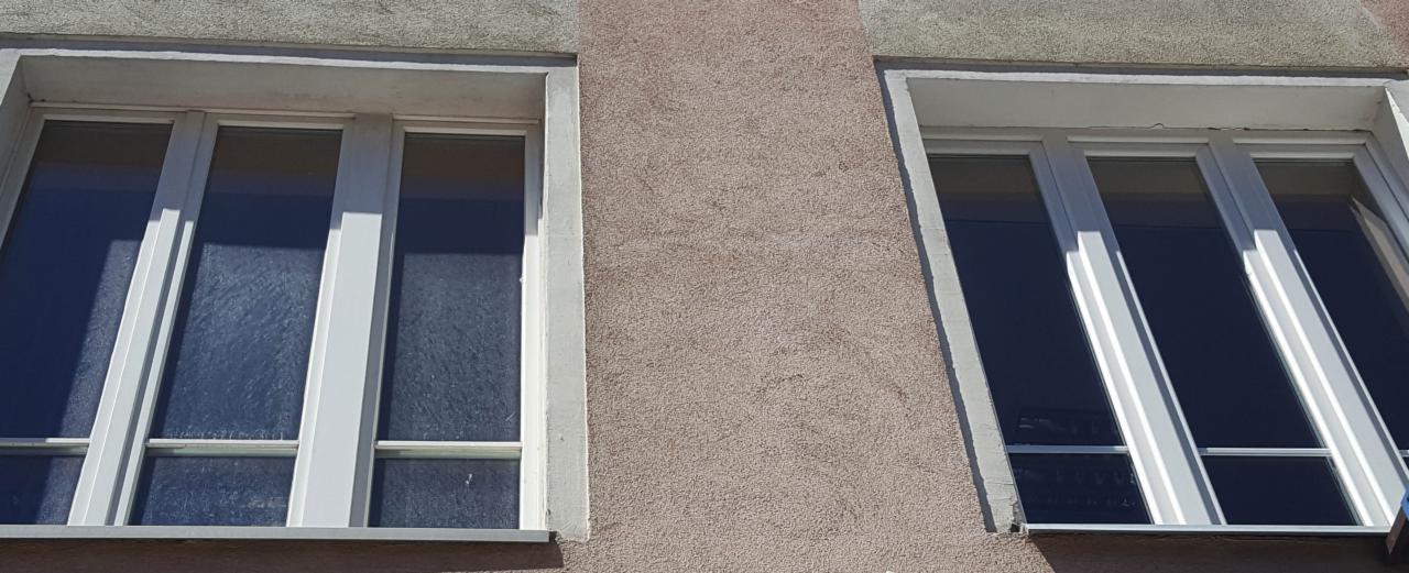 Dwa okna z białymi ramami na elewacji budynku o beżowej, chropowatej strukturze tynku. Na szybach widoczne smugi, sugerujące niedawne mycie.