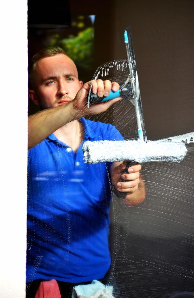 Mężczyzna w niebieskiej koszulce, z widoczną bródką i niebieskimi oczami, myje okno za pomocą ściągaczki z niebieskim uchwytem, pozostawiając smugi piany na szkle.