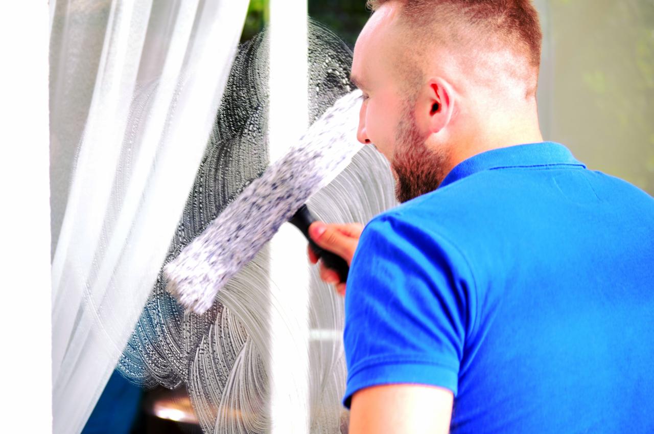 Mężczyzna w niebieskiej koszulce polo myje okno za pomocą ściągaczki, widoczne smugi wody i mydlin na szybie.