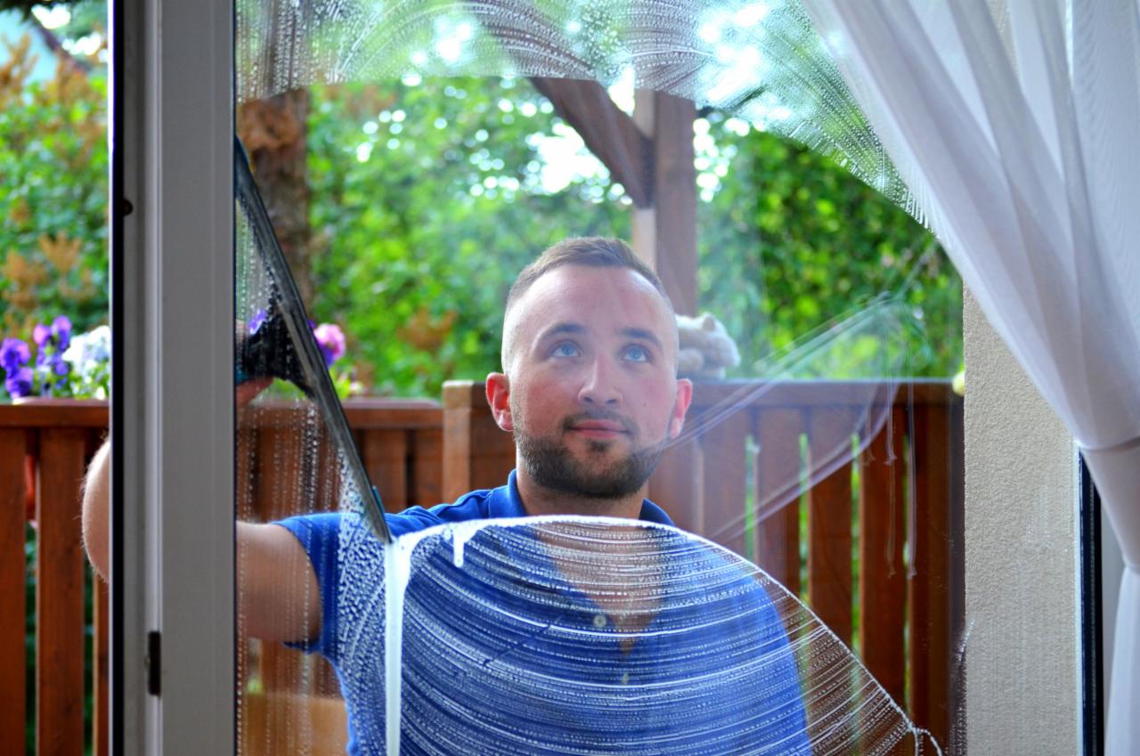 Mężczyzna z krótką brodą czyści okno za pomocą ściągaczki, widoczne smugi detergentu na szybie, w tle balkon z kwiatami i zieleń.