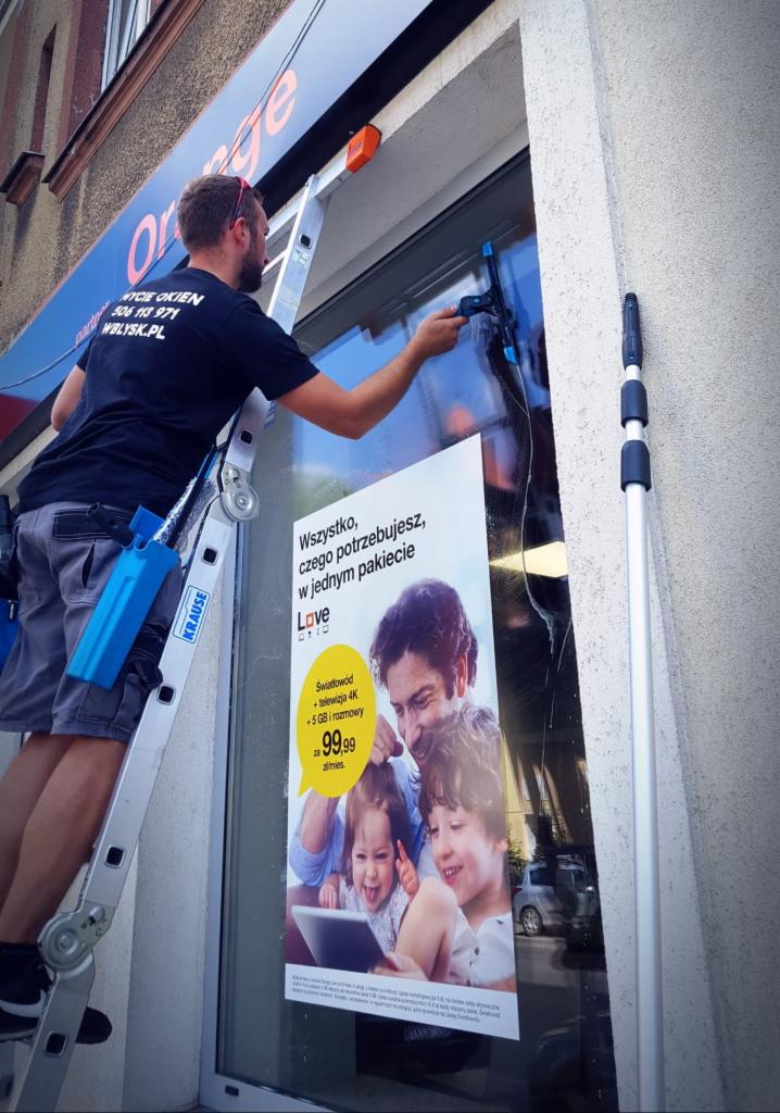 Pracownik myje okno na wysokości przy użyciu ściągaczki, stojąc na drabinie Krause. Na oknie naklejony plakat reklamowy sieci Orange.