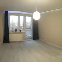 Pusty pok&oacute;j po remoncie z szarymi panelami podłogowymi, beżowymi ścianami, grzejnikiem pod oknem i wiszącą lampą w kształcie kuli, w tle widok na osiedle z okna.