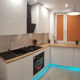 Nowoczesna kuchnia w bieli z drewnianym blatem, czarnym zlewem i płytą gazową, podświetleniem LED w kolorze turkusowym pod szafkami i pomarańczowymi roletami w oknie.