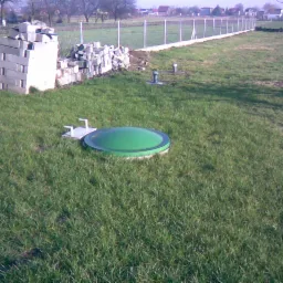 Przydomowa przepompownia ścieków z zieloną pokrywą w trawie, widoczne betonowe bloczki i elementy instalacji, w tle ogrodzenie i zabudowania.