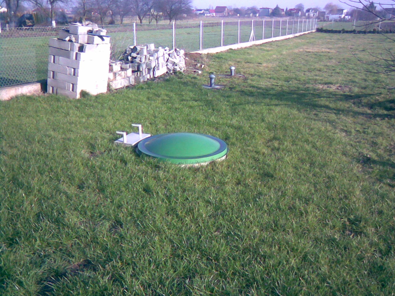 Przydomowa przepompownia ścieków z zieloną pokrywą w trawie, widoczne betonowe bloczki i elementy instalacji, w tle ogrodzenie i zabudowania.