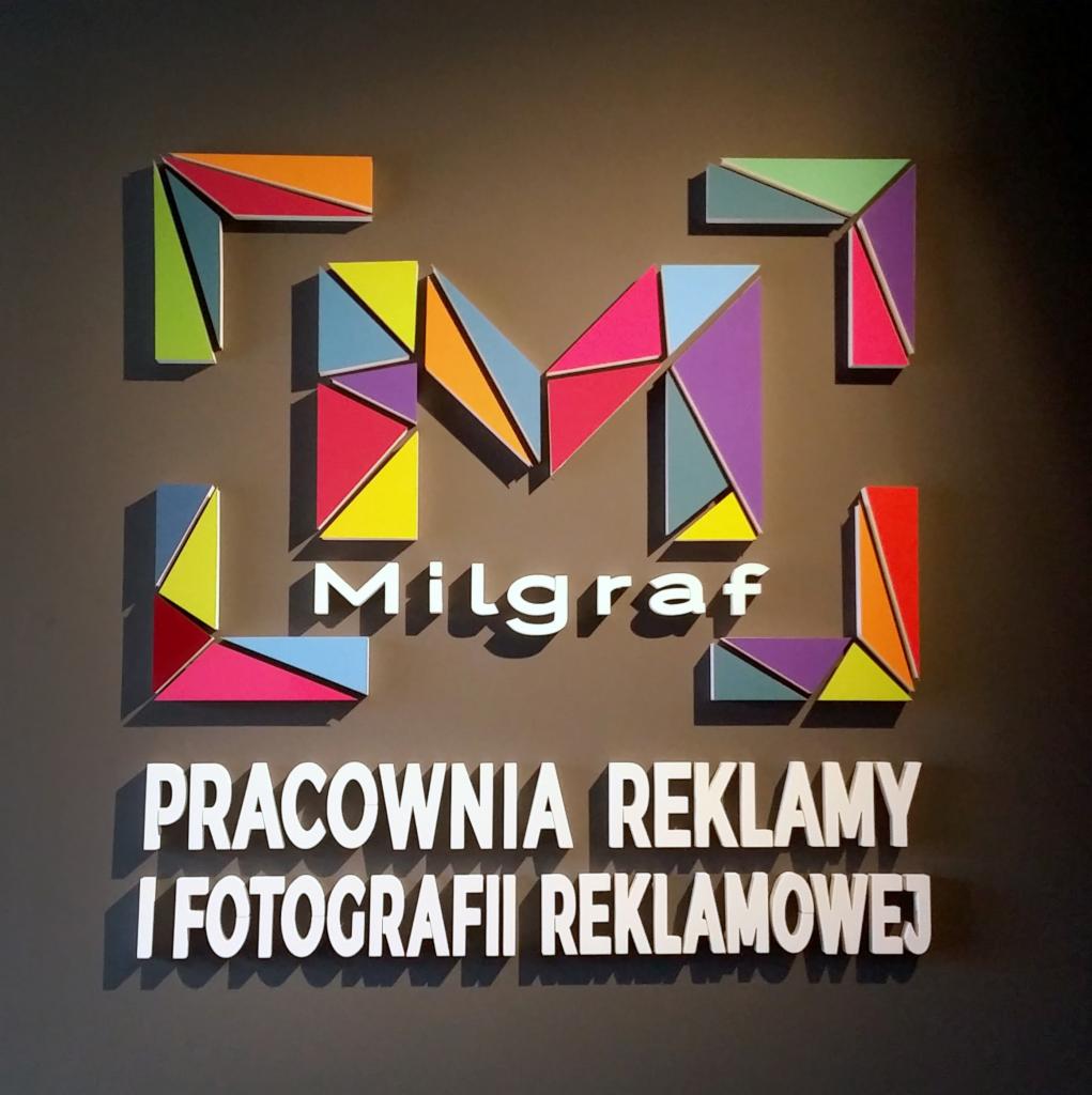 Ścienna tablica informacyjna z logo firmy Milgraf, pracowni reklamy i fotografii reklamowej. Logo składa się z wielobarwnych trójkątnych elementów tworzących literę M.