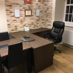 Biuro z brązowym biurkiem na wymiar w kształcie litery L, czarnymi krzesłami, skórzanym fotelem obrotowym i ceglaną ścianą z dyplomami.
