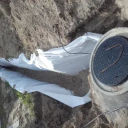 Wykop pod drenaż z ułożoną geowłókniną i zasypany żwirem, widoczny betonowy krąg z żeliwnym włazem oraz wystająca ziemia.